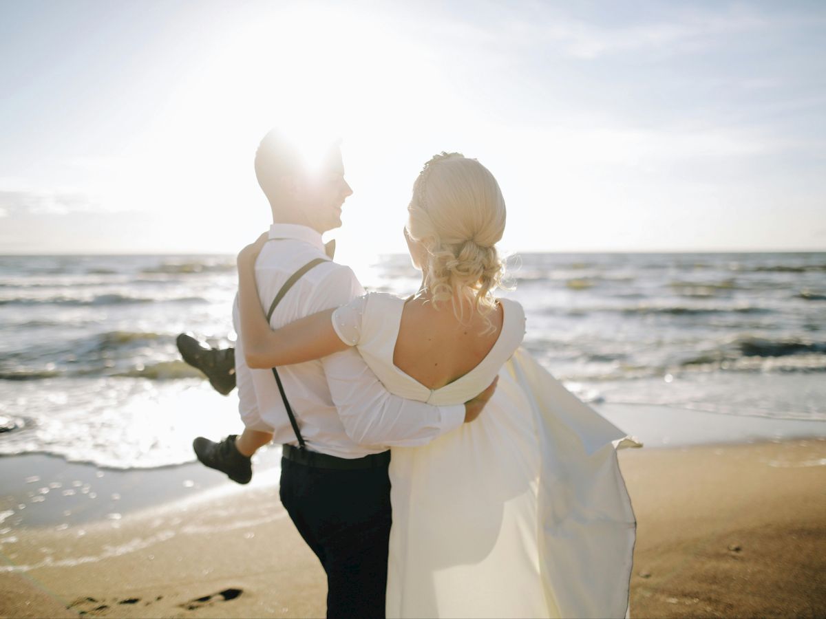 A couple is on a beach; the man carries the woman as the sun sets over the ocean.