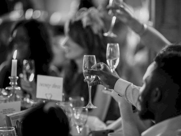 People are raising glasses in a celebratory toast at an indoor event. The scene is in black and white, creating an elegant atmosphere.