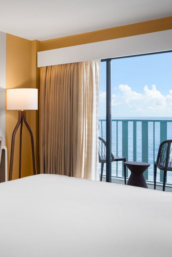 A cozy hotel room features a bed, floor lamp, and a balcony with chairs overlooking a scenic ocean view under a clear blue sky.