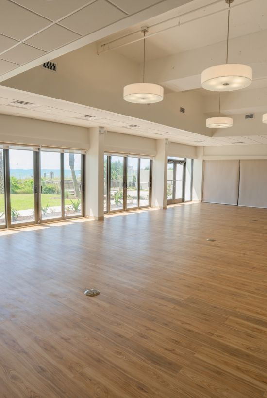 This image shows a spacious, sunny room with wooden floors, large windows overlooking greenery, and modern hanging lights on the ceiling.