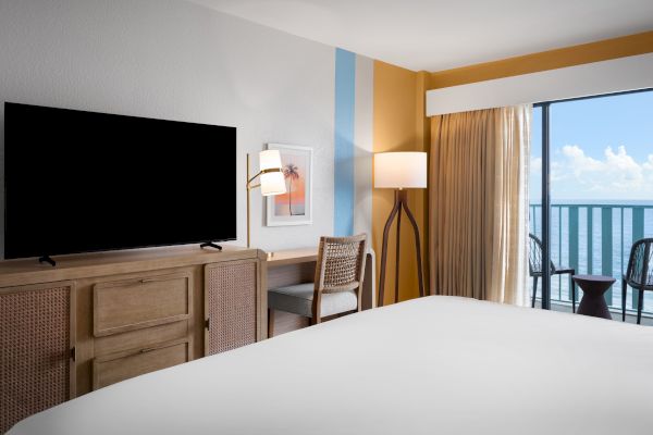 The image shows a hotel room with a bed, TV, desk, chair, a lamp, and a balcony with an ocean view under a blue sky and clouds.