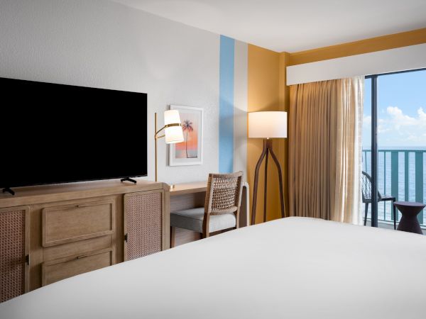 The image shows a hotel room with a bed, TV, desk, chair, a lamp, and a balcony with an ocean view under a blue sky and clouds.