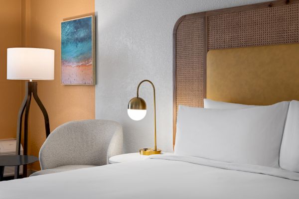 A cozy hotel room with a bed, modern lamp, white pillow, chair, wall art, and a stylish floor lamp.