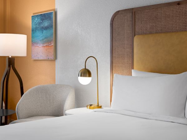 A cozy hotel room with a bed, modern lamp, white pillow, chair, wall art, and a stylish floor lamp.