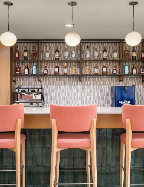 A modern bar setup with pink chairs, pendant lights, shelves with bottles, and a coffee machine on the counter.