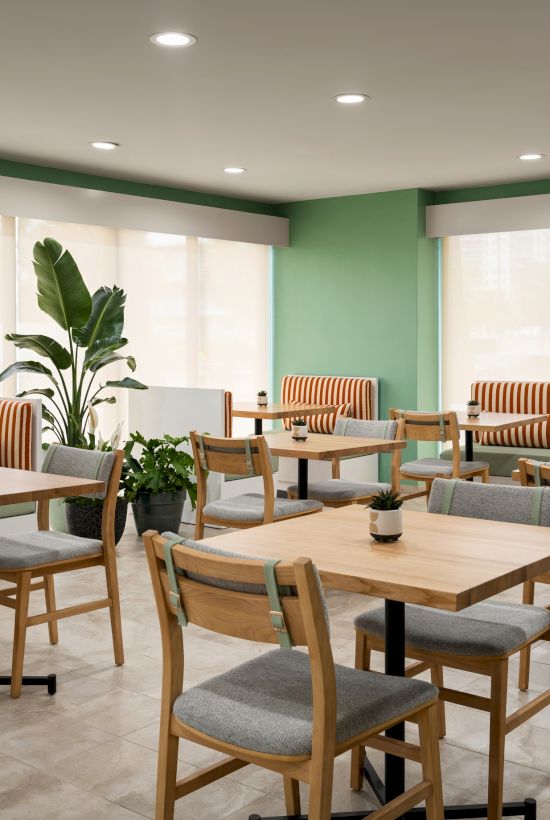 A cozy cafe with wooden tables, chairs, and striped benches; potted plants add greenery to the sunlit space.