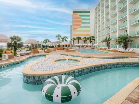 This image shows a hotel pool area with a lazy river, surrounded by sun loungers, umbrellas, and a mural on the building wall.