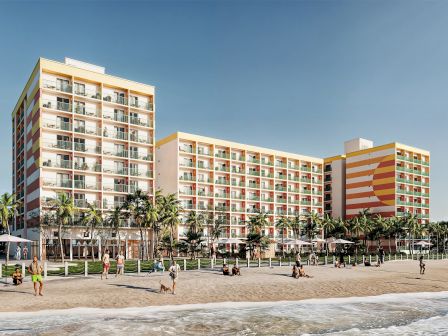 The image shows a colorful beachfront hotel with people on the sandy beach and palm trees lining the perimeter, under a clear sky.
