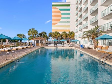 A sunny hotel pool with lounge chairs, teal umbrellas, palm trees, and a colorful building tower beside it.
