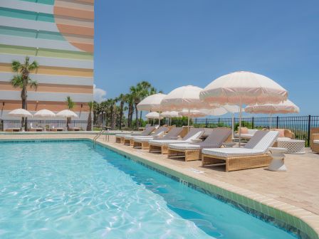 A sunny poolside scene with lounge chairs, umbrellas, and a colorful building in the background, set against a clear blue sky.
