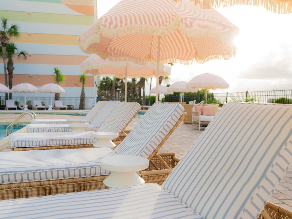 Poolside scene with striped lounge chairs, pink umbrellas, and a pastel-colored hotel in the background under a sunny sky.