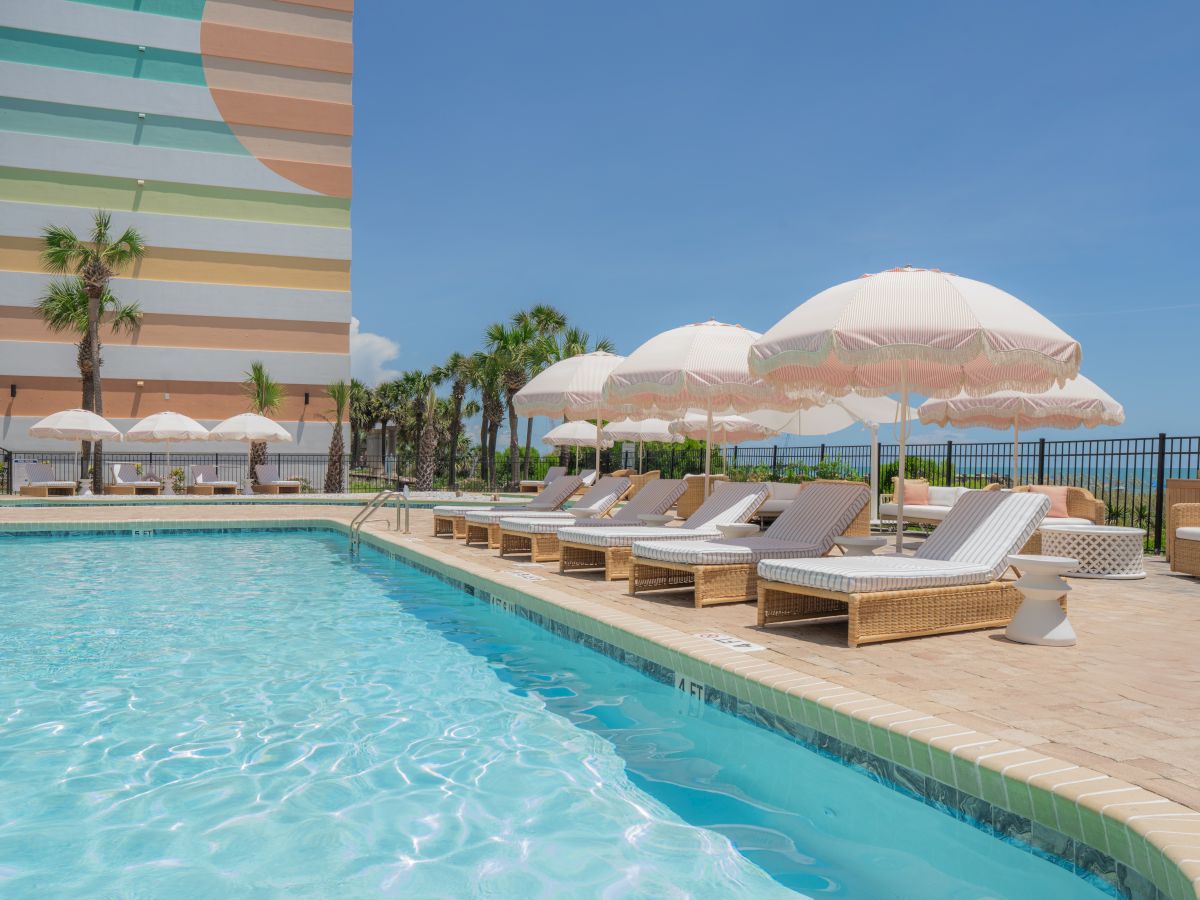 A sunny poolside scene with wicker lounge chairs, white umbrellas, and a colorful building, surrounded by palm trees under a clear blue sky.