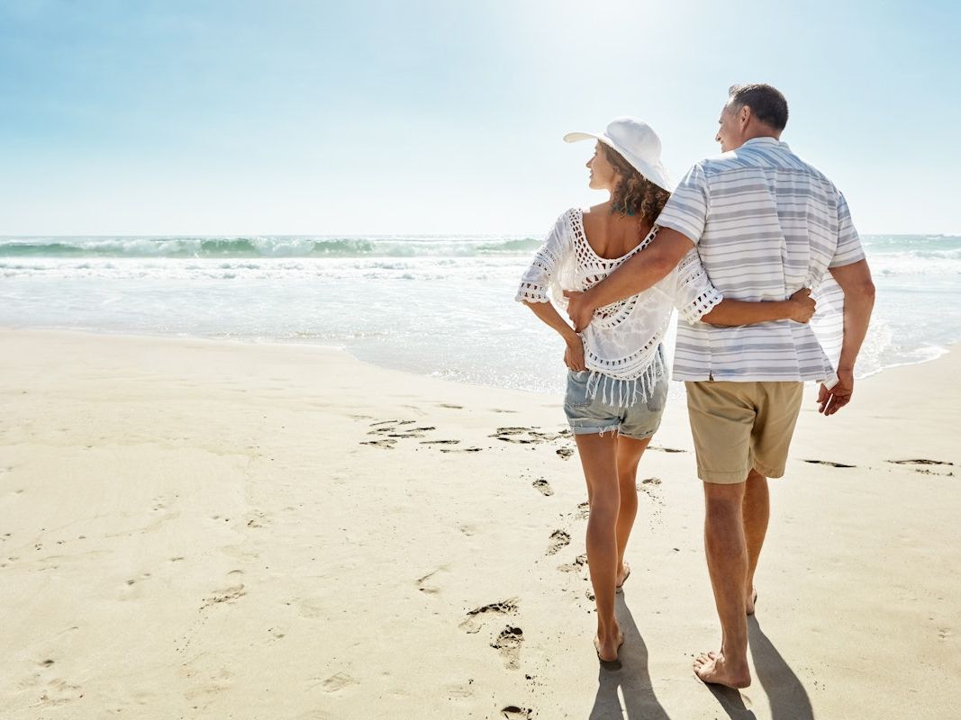 A couple in summer clothes strolls along a sunny beach, leaving footprints by the waves.