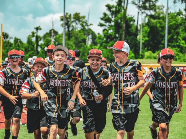 A group of kids, wearing "Hornets" jerseys and caps, joyfully run on a sports field, surrounded by greenery and colorful banners.