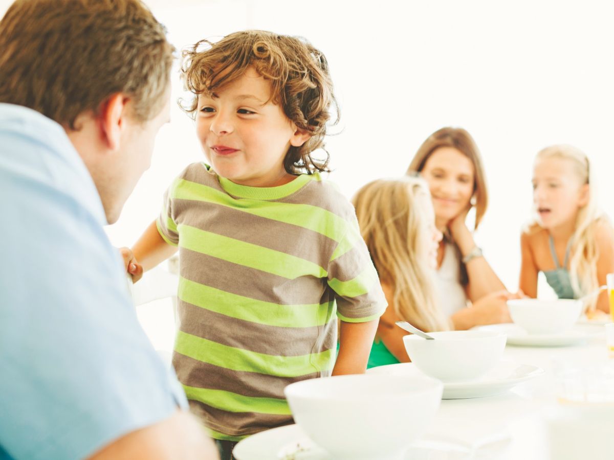 A family meal at a bright table: a smiling child in a striped shirt interacts with a man, while others watch, bowls and cups nearby, in a warm, cheerful scene.