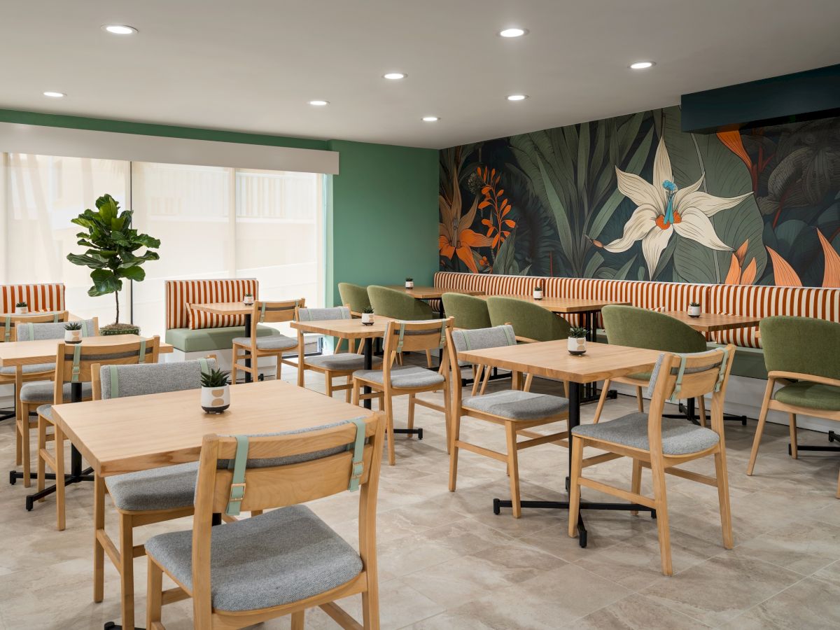The image shows a modern, cozy dining area with wooden tables and chairs, green accents, and a floral mural on the wall.