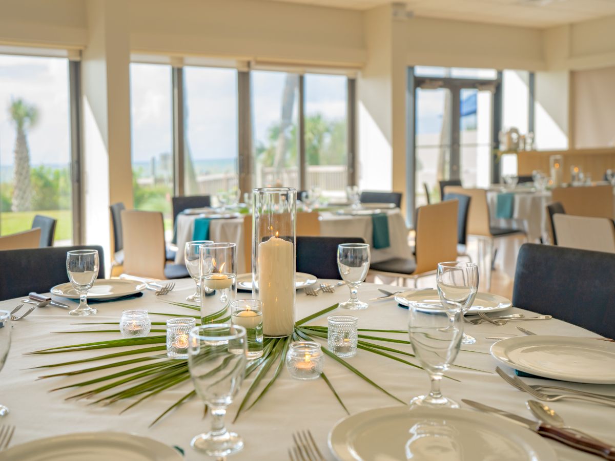 A dining area with elegantly set tables, glassware, white plates, and palm leaf centerpieces, surrounded by large windows and chairs.