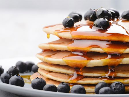 A stack of pancakes topped with blueberries and drizzled with syrup on a plate, surrounded by more blueberries.