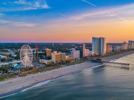 A coastal cityscape at sunset with a Ferris wheel, tall buildings, a sandy beach, and a pier stretching into the calm ocean waters.