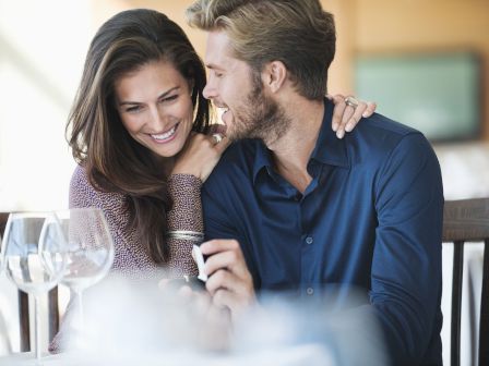 A couple sits closely at a table, smiling and sharing an intimate moment, with their surroundings suggesting a restaurant setting.