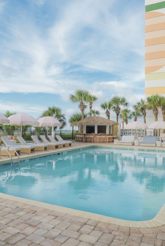 A sunny hotel pool area with lounge chairs, palm trees, and a colorful striped building in the background, under a blue sky.