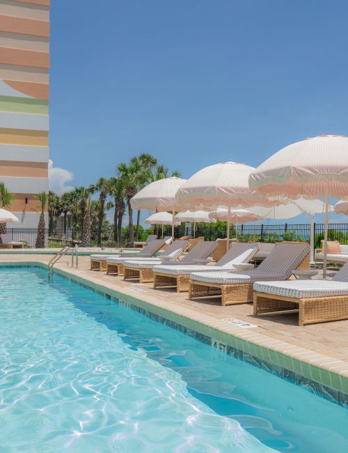 A poolside area with wicker lounge chairs, white umbrellas, surrounded by a modern building and palm trees under a clear blue sky.