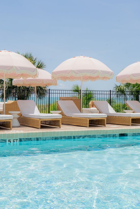 Poolside scene with clear water, lounge chairs, and pink umbrellas under a blue sky, creating a relaxing outdoor setting.