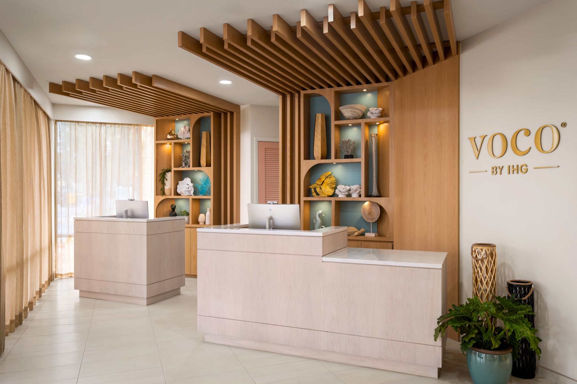 The image shows a modern hotel reception desk with wood accents, decorative shelves, and a plant, branded with "VOCO by IHG."