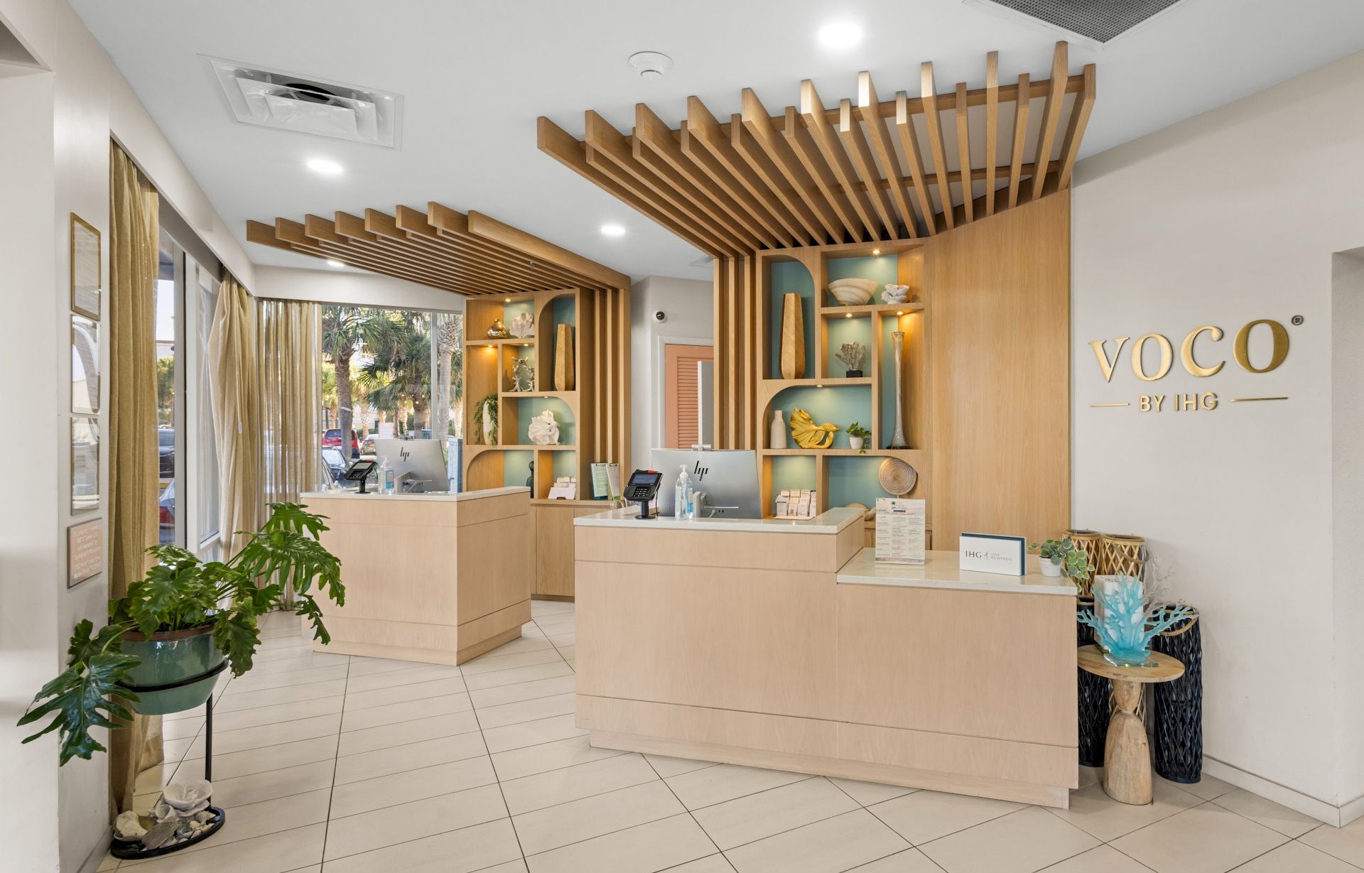 Modern reception area for voco by IHG, with light wood slatted ceilings and counters, decor shelves, and plants.