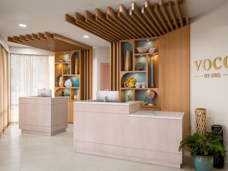 A modern hotel reception area featuring wooden decor, two desks, decorative shelves, and a welcoming atmosphere with "VOCO by IHG" branding.