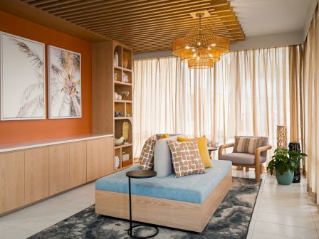 A cozy living room with a modern sofa, artwork, a rug, indoor plants, wooden ceiling, and large windows with curtains.