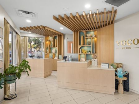 A modern hotel reception area for &ldquo;voco by IHG,&rdquo; with light wood counters, shelves, plants, and bright ceiling lights.