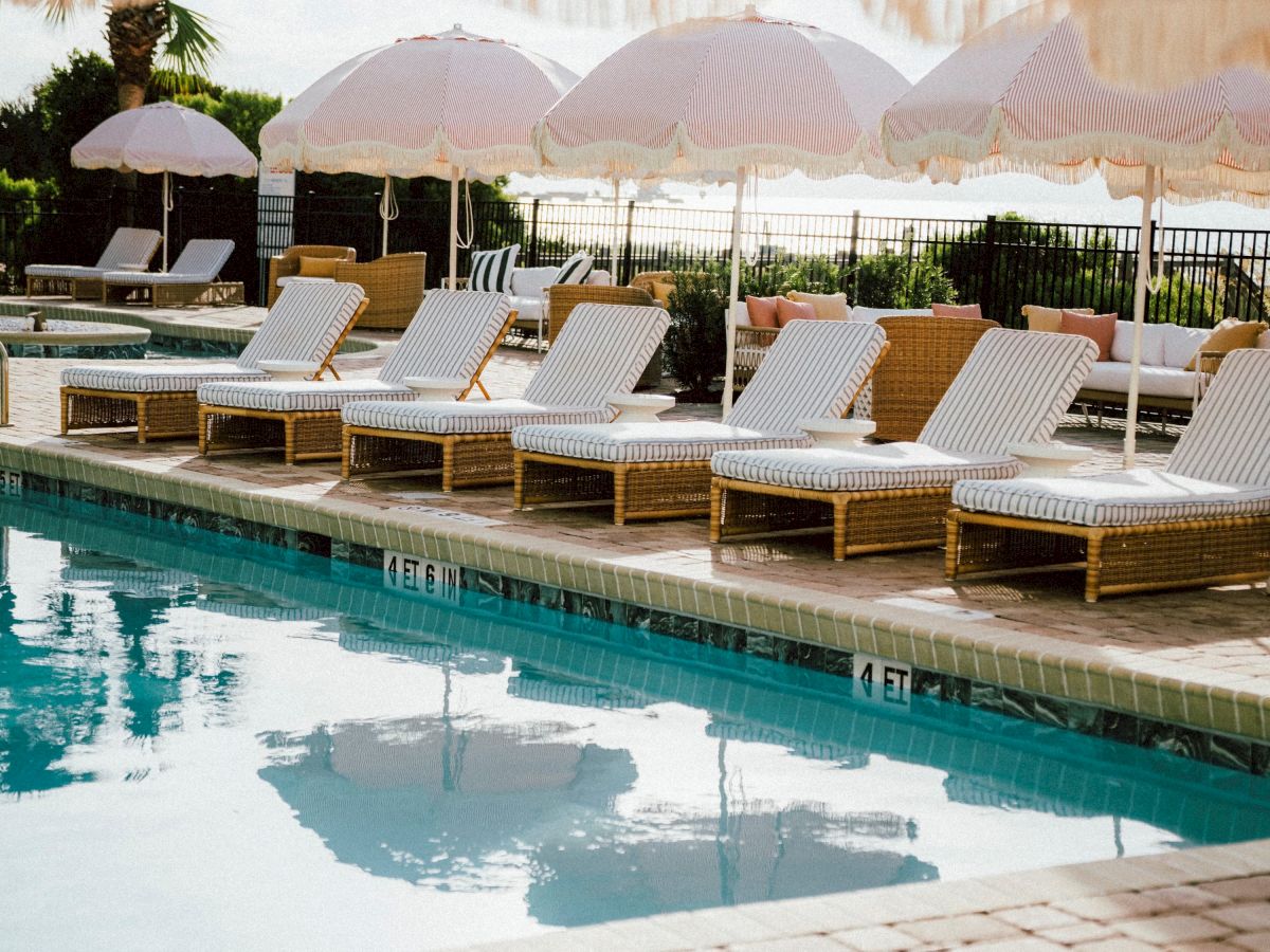 A row of white lounge chairs and umbrellas by a poolside, with a striped awning overhead, set near a scenic body of water.