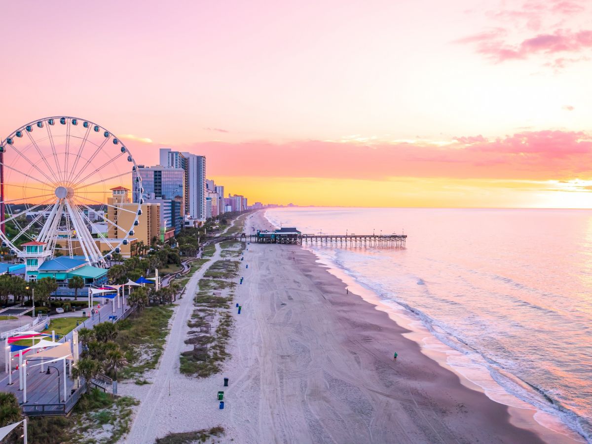 A coastal cityscape at sunset with a Ferris wheel, high-rise buildings, a sandy beach, and a calm ocean stretching to the horizon.