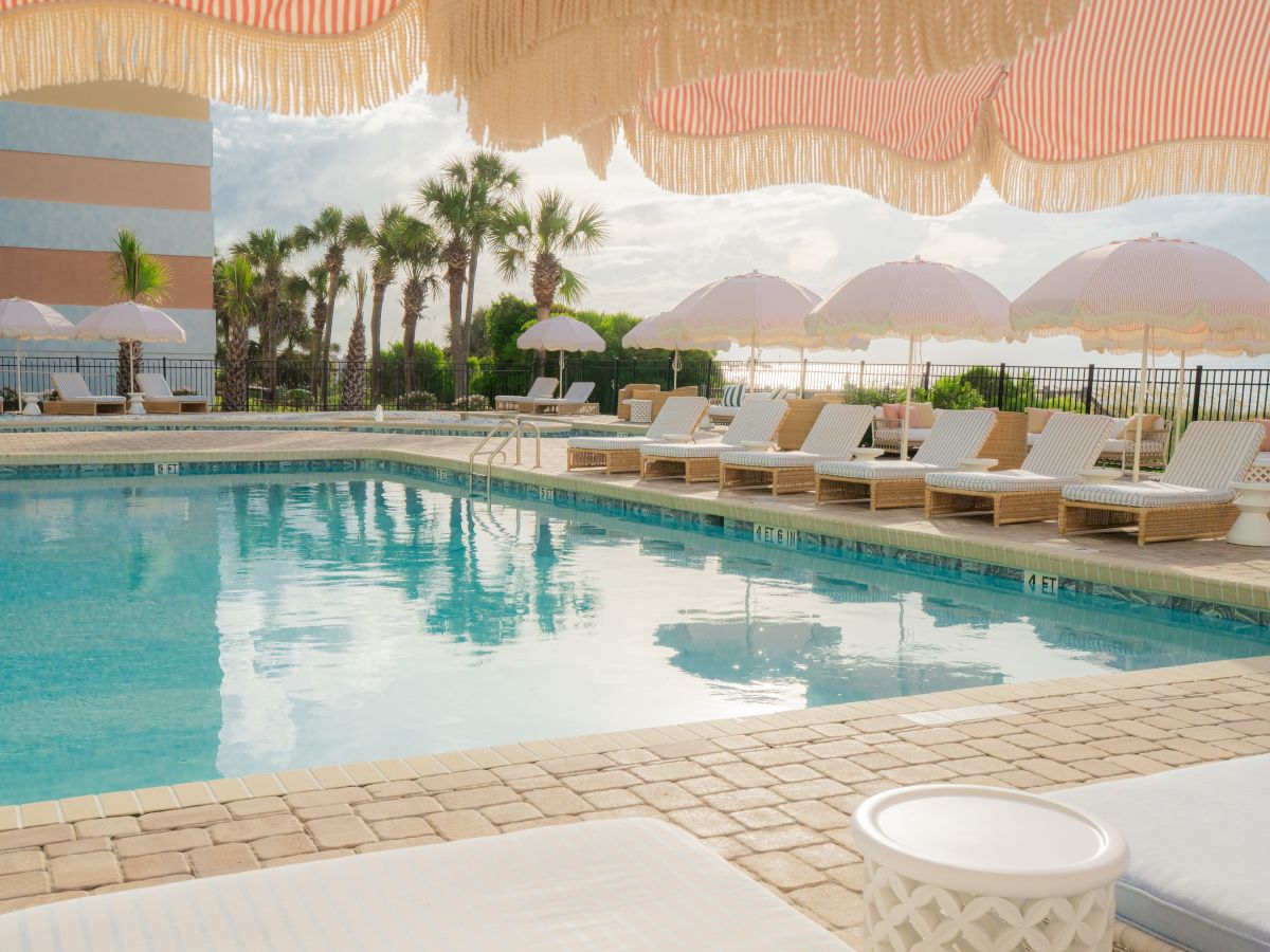 A serene poolside scene with sun loungers, pink umbrellas, and palm trees under a sunny sky, perfect for a relaxing day.