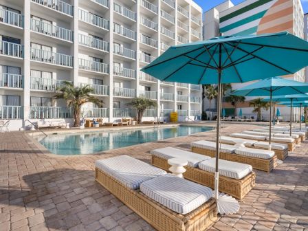 A sunny hotel poolside with multiple lounge beds, teal umbrellas, and a swimming pool in front of white apartment buildings.