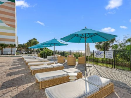 A sunny pool deck with cushioned lounge chairs, green umbrellas, and safety signs behind a metal fence.