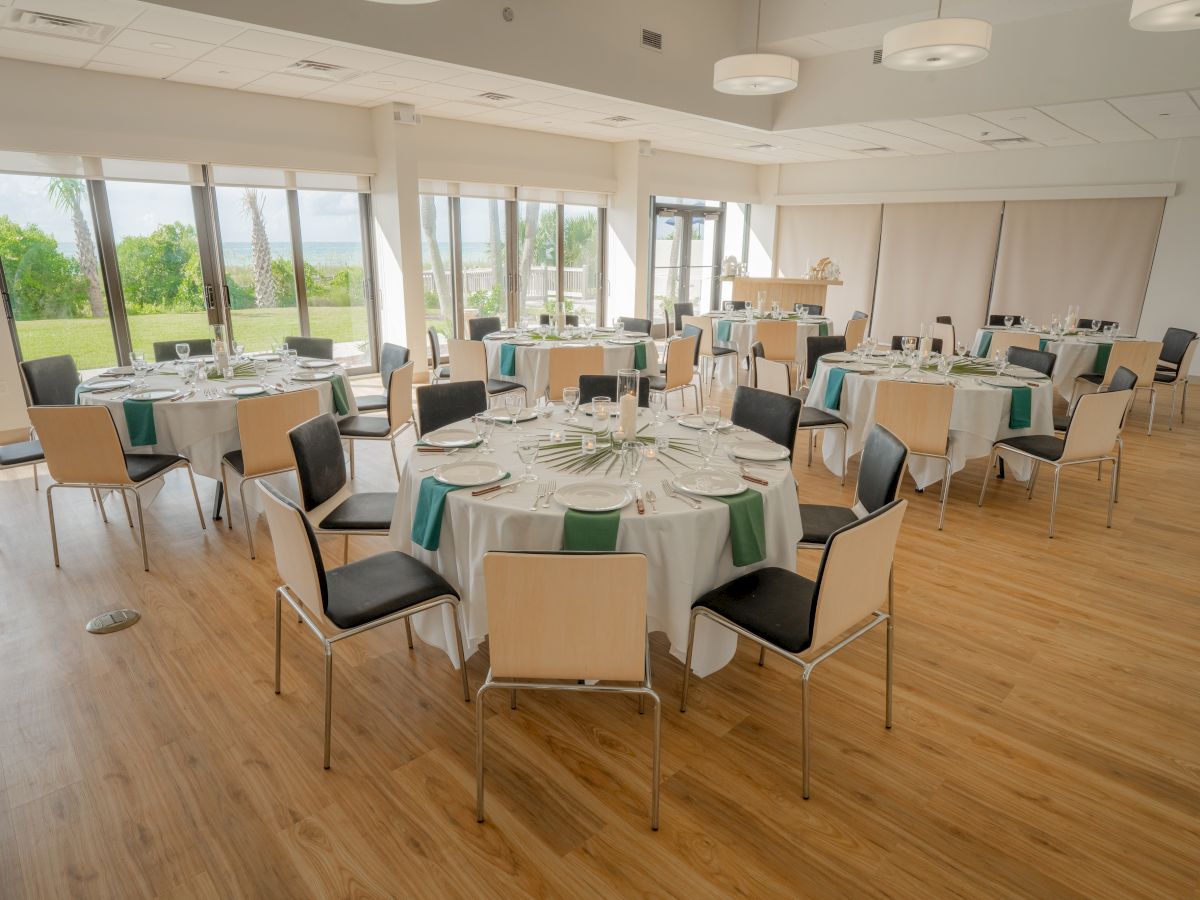 The image shows a banquet hall with round tables set for dining, including white tablecloths, plates, and chairs on a wooden floor.
