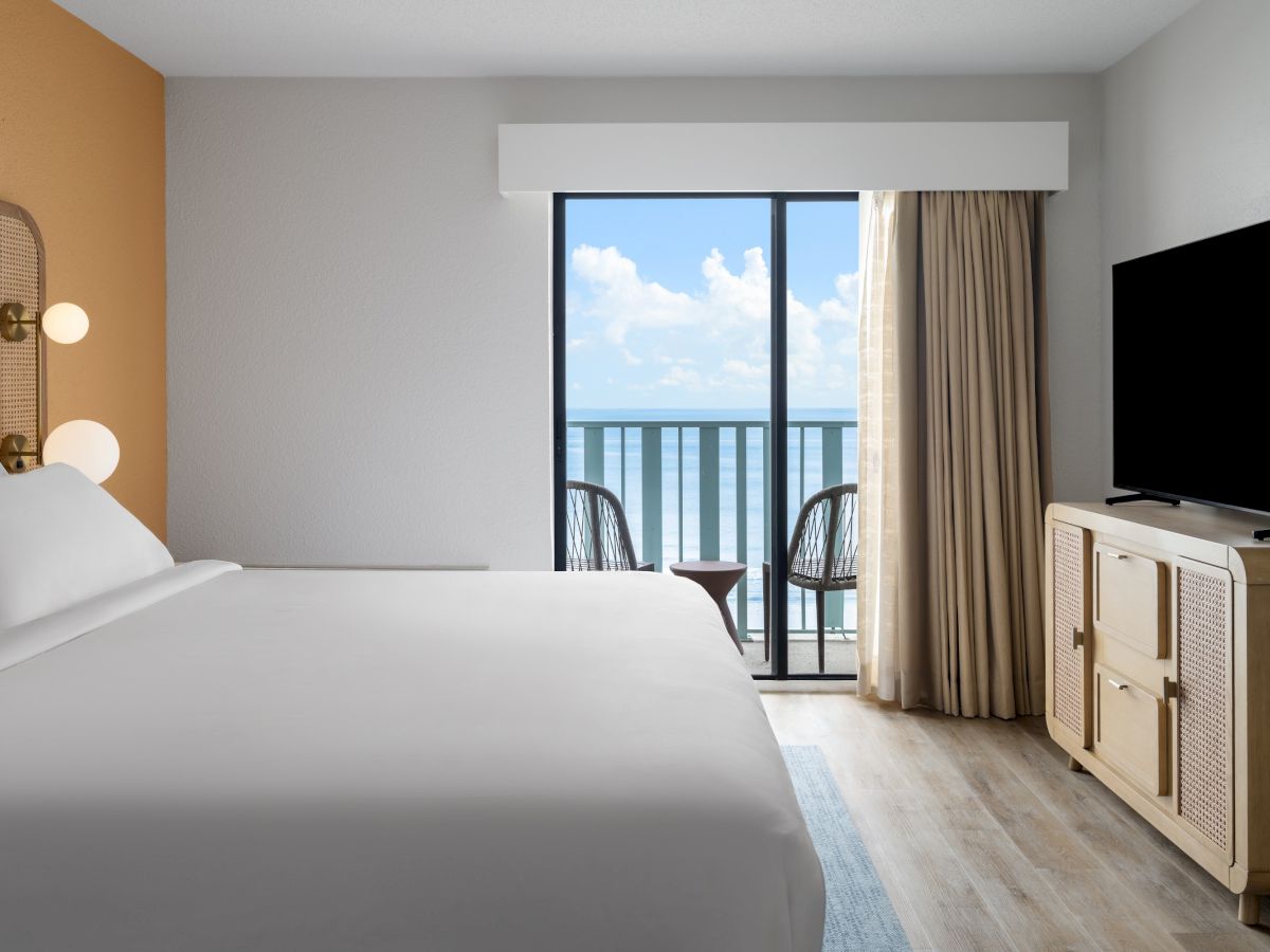A hotel room with a bed, flat-screen TV, and a window showing a balcony with chairs and a view of the ocean under a blue sky.