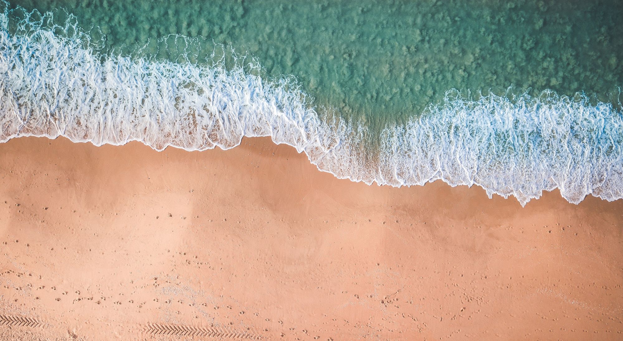 Aerial view of a sandy beach meeting turquoise waves, foamy white surf washing onto shore, clear water transitioning from deep to shallow.