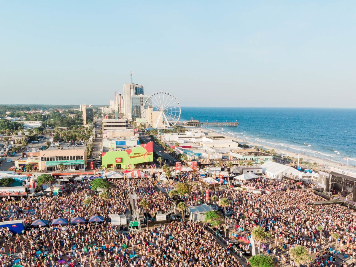 A massive outdoor festival on a sunny beach boardwalk, with a crowd filling the coast, rides, stages, and a ferris wheel in the background.