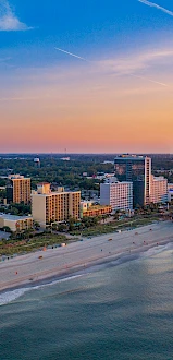 A coastal cityscape at sunset with a beach, high-rise buildings, a Ferris wheel, and a pier extending into the ocean, under a colorful sky.