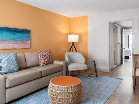 The image shows a modern living room with a sofa, chair, lamp, artwork, and a round table on a rug, next to an open doorway.