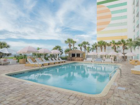 A sunny hotel pool area with lounge chairs, palm trees, umbrellas, and a colorful building in the background by the sea.