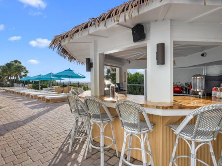 An outdoor poolside bar with thatched roof, metal chairs, umbrellas, and a drinks prep area under a sunny blue sky.
