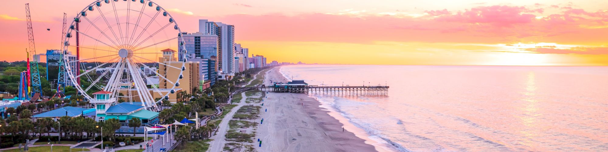 A sunny beach scene at sunset with a ferris wheel, hotels, and a pier along a calm shoreline.