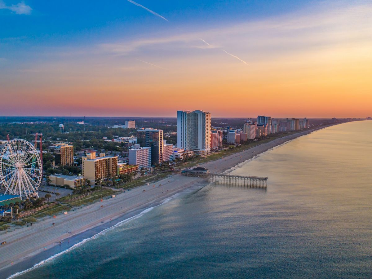 A sunny coastal city with a Ferris wheel, beachfront hotels, a long pier, and a calm ocean sunset over high-rise buildings along a golden sandy shore.