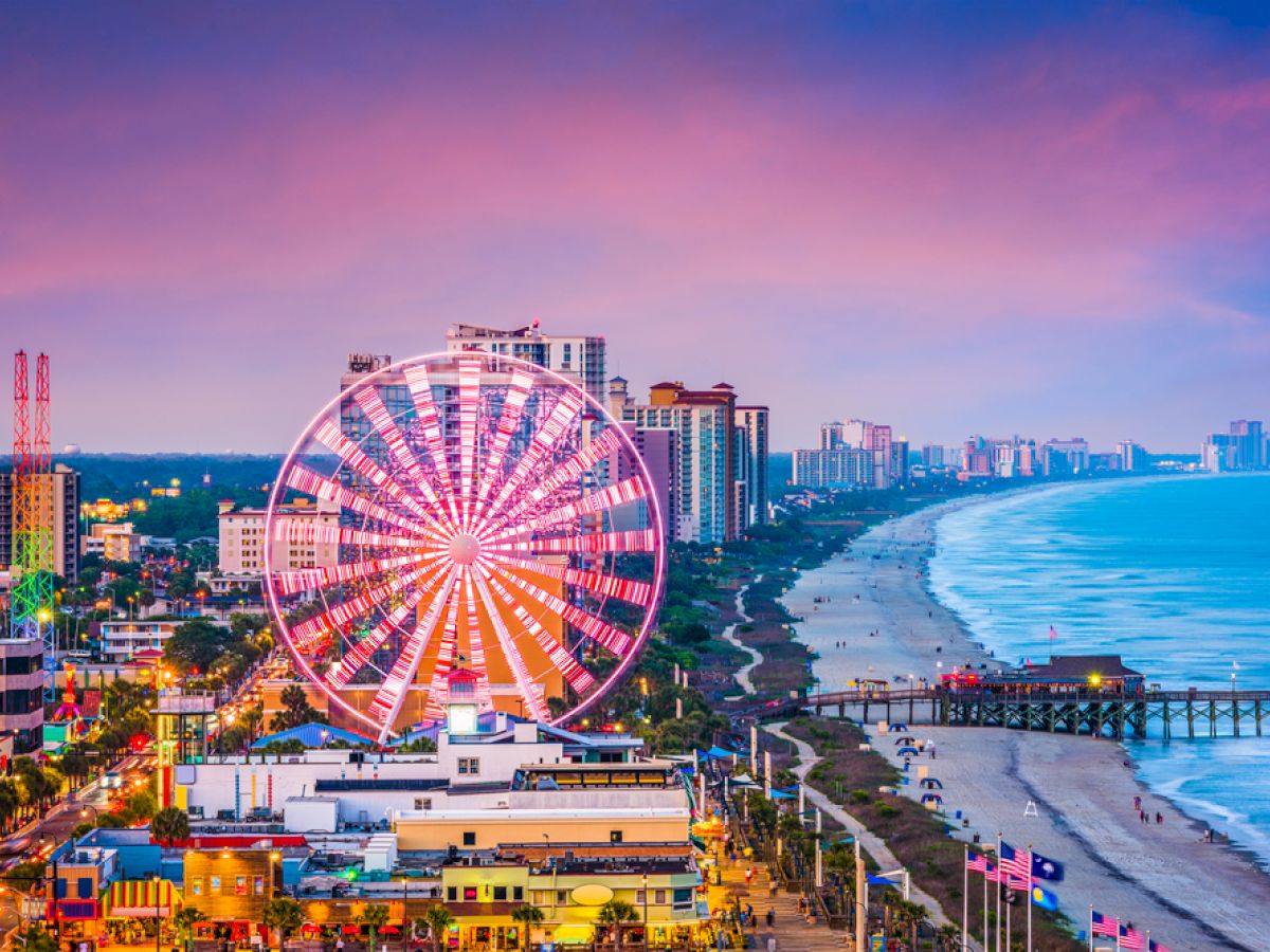 A bright Ferris wheel along a busy beachfront boardwalk at sunset, with colorful buildings and a long pier extending into the ocean. (140 chars)