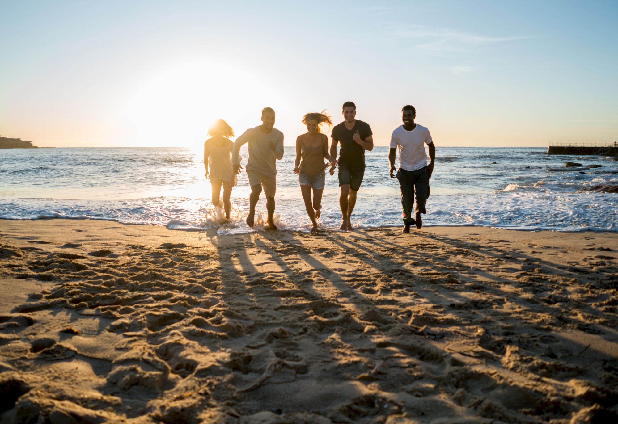 A group of five men walking along a sunny beach at sunset, casting long shadows on the sandy shore near the water&rsquo;s edge. (ends sentence)