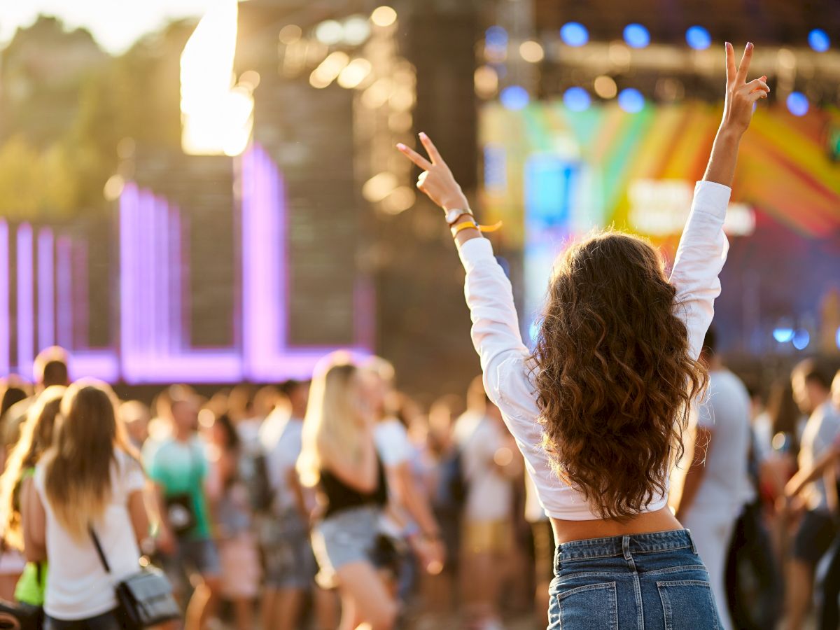 A woman with arms raised at a crowded outdoor concert or festival, stage lights glow in the background, everyone dancing and enjoying the music.